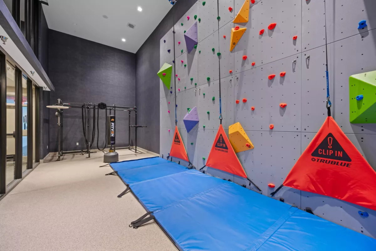 Indoor rock climbing wall with colorful holds, blue mats, and belay systems attached.
