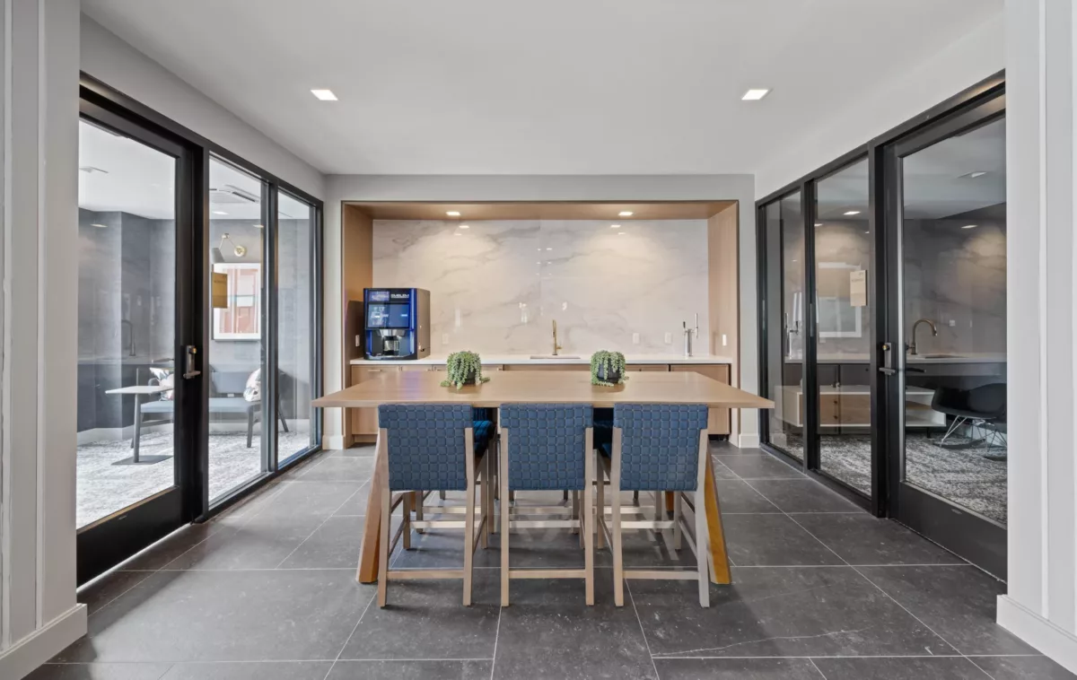 Coffee bar with a wood table, blue chairs, a coffee machine, and glass doors leading to seating areas.