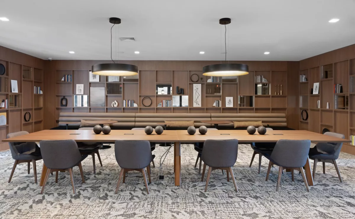 Library-style meeting room with long wood table, grey chairs, wall shelves, and black pendant lights.