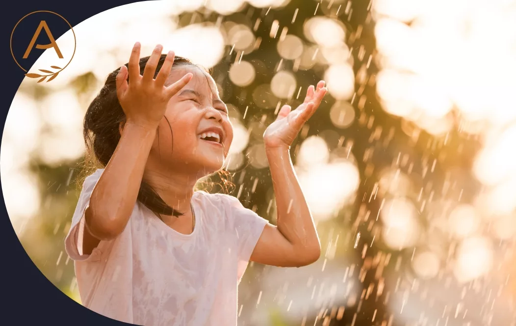 Smiling child with arms raised, playing in falling water under warm sunlight with blurred foliage in the background.