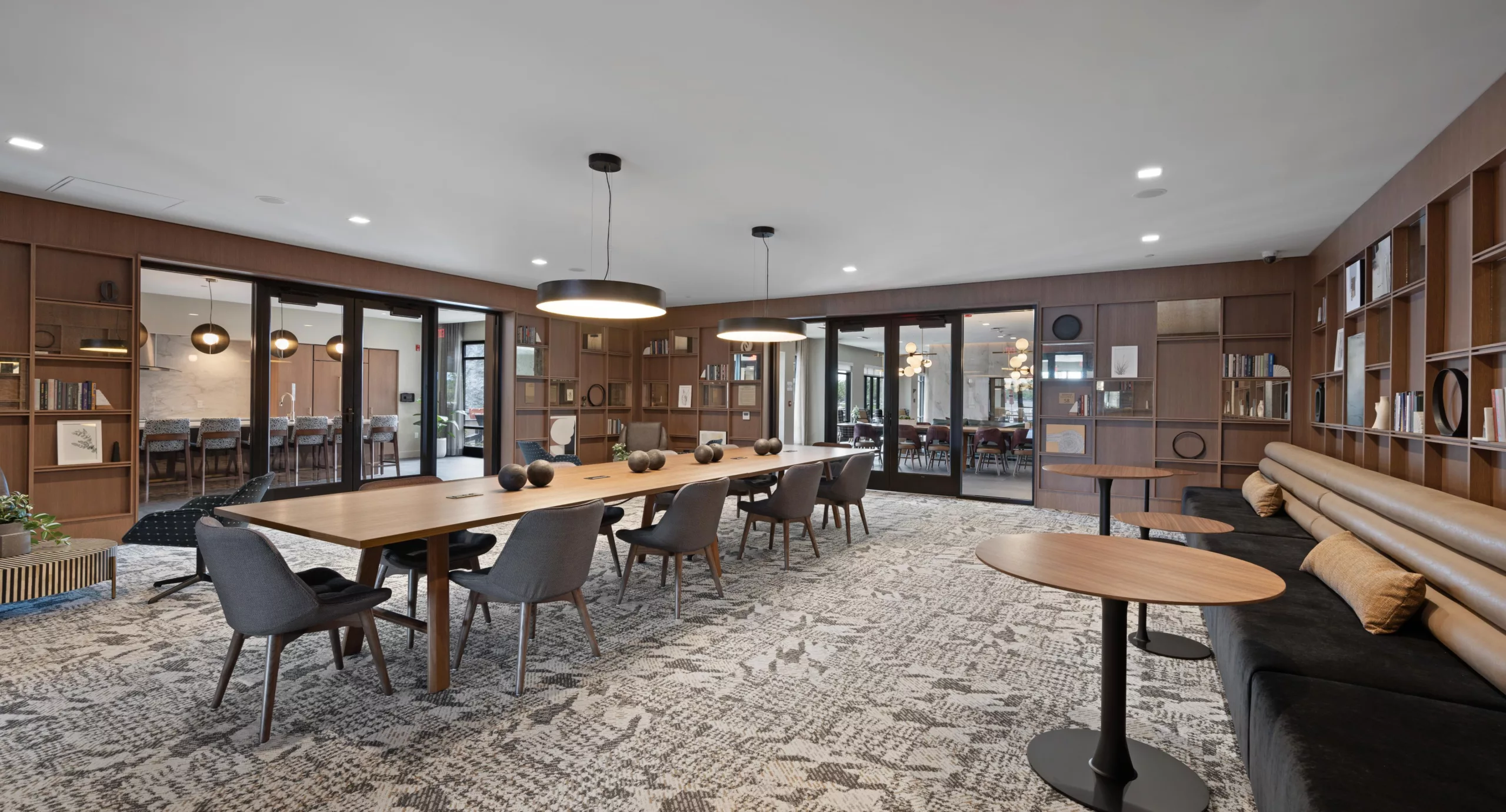 Spacious meeting room with wood table, grey chairs, bookshelves, and glass doors leading to adjacent rooms.